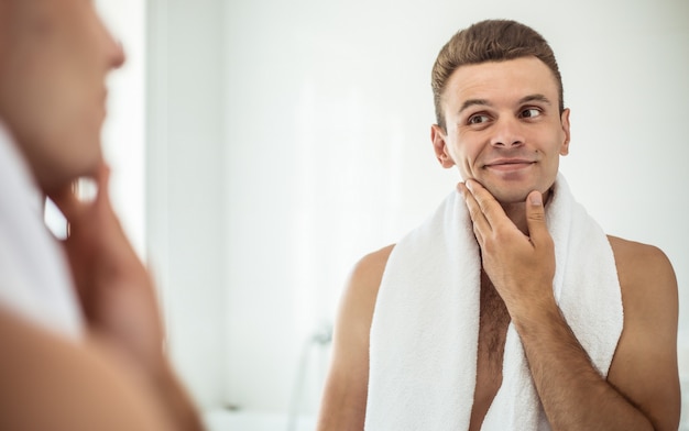 Красивый молодой человек бреет бороду в ванной комнате. Портрет стильного обнаженного бородатого мужчины, разглядывающего свое лицо в домашнем зеркале.