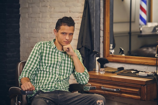 Handsome young man in shirt and jeans looking at the camera and sitting in a chair at the barbershop. He looks confident