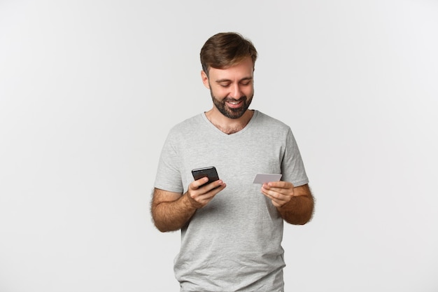 Handsome young man shopping online, holding credit card and mobile phone, standing over white