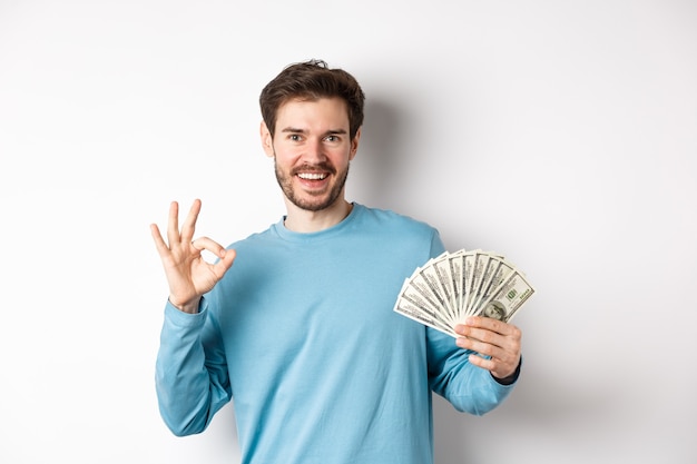 Handsome young man showing quick loans money, make okay gesture and smiling with cash, standing over white background.