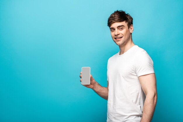 Handsome young man showing smartphone with blank screen and smiling at camera on blue background
