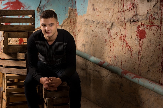 Handsome young man sitting on empty packing crates in terrible basement in a Halloween horror concept