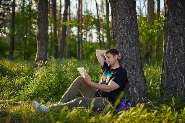 Handsome young man sitting in the forest on the grass, leaning against a tree, reading a book