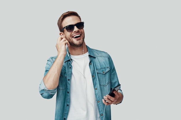 Handsome young man smiling and listening music while standing against grey background