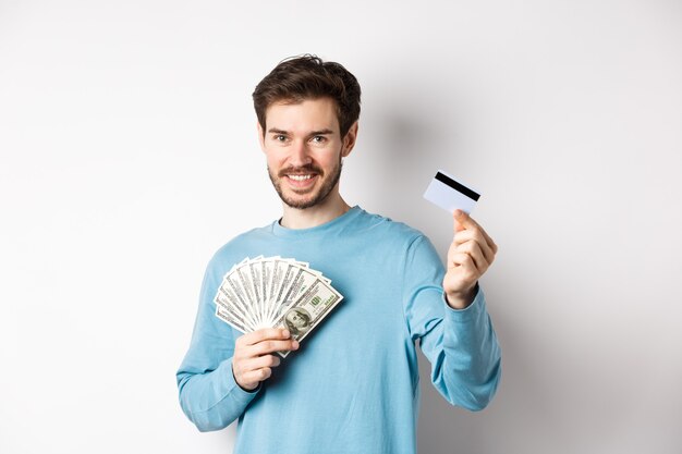 Handsome young man smiling and offering payment in cash and contactless, showing money with plastic credit card, standing on white background.