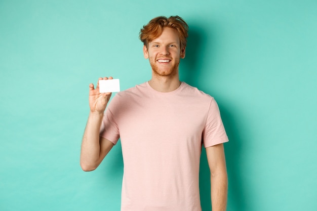 Handsome young man smiling and showing plastic credit card, standing in t-shirt against turquoise background.