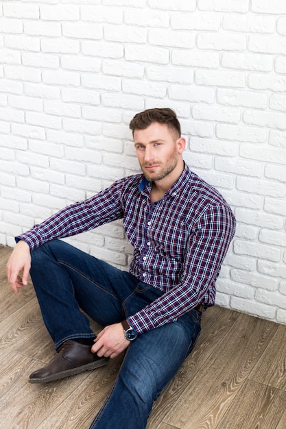 Handsome young man smiling while sitting on the floor and leaning at the wall