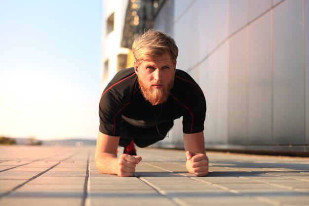 Handsome young man in sports clothing keeping plank position while exercising outdoors