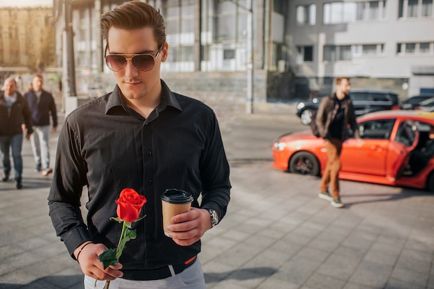 Handsome young man stand outside. He holds red rose and cup of drink. Guy wears sunglasses. There are people and cars behind him.
