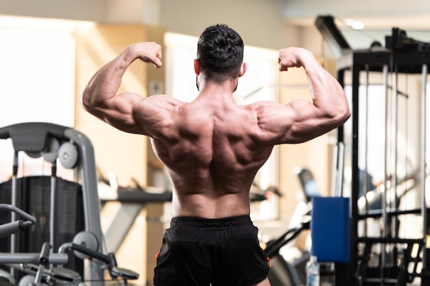 Photo handsome young man standing strong in the gym and flexing muscles muscular athletic bodybuilder fitness model posing after exercises