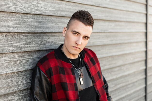 Handsome young man in a stylish plaid jacket with a trendy hairstyle in a black trendy t-shirt is resting near a wooden wall on a spring day