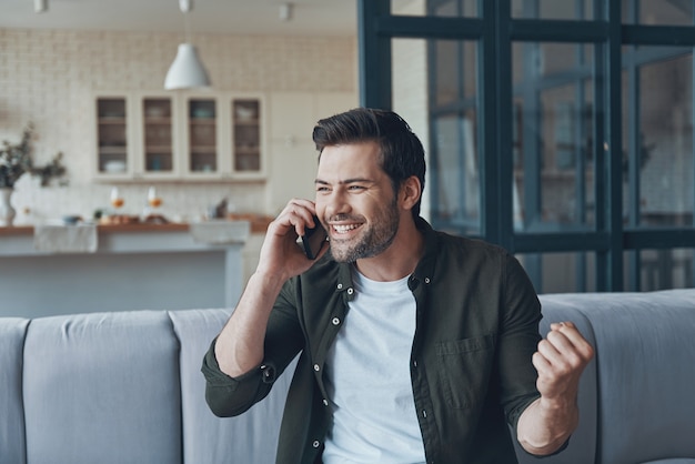 Handsome young man talking on smart phone and looking away while sitting on the sofa at home