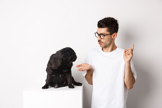 Handsome young man teaching dog commands, talking to cute black pug, standing over white background