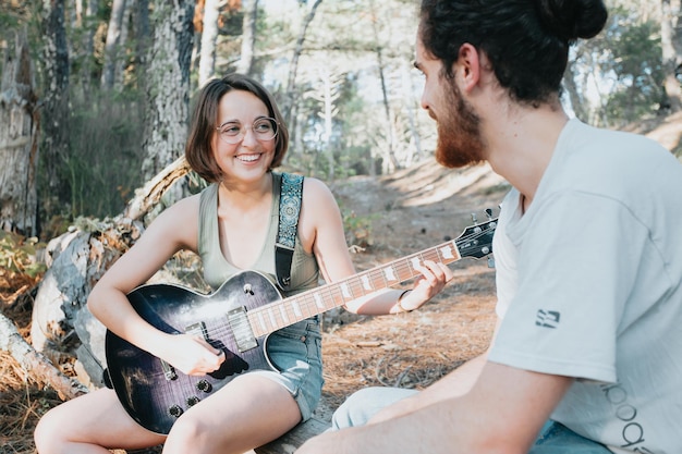 Handsome Young Man Teaching hipster Girl to Play Guitar on the forest trees during a sunset. Learning a new skill joyful concept. Music with copy space. Young couple electric guitar