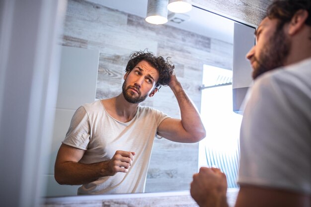 Handsome young man touching his hair with hand and grooming in bathroom at home. White metrosexual man worried for hair loss and looking at mirror his receding hairline.