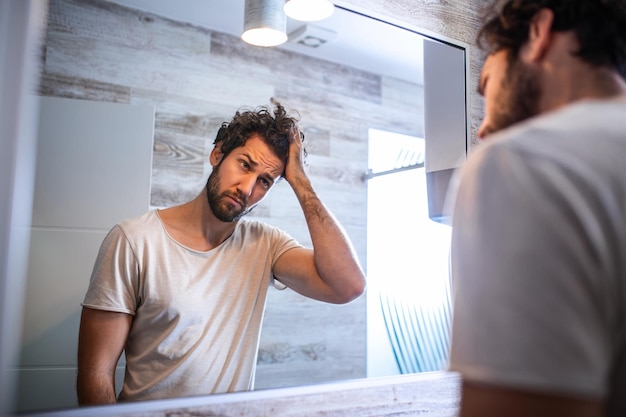 Handsome young man touching his hair with hand and grooming in bathroom at home. White metrosexual man worried for hair loss and looking at mirror his receding hairline.