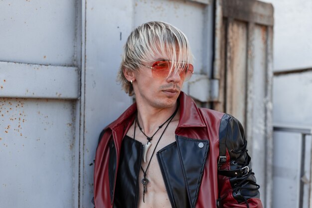 Handsome young man in trendy black and red leather jacket in red sunglasses with stylish hairstyle outdoors near the metallic gray wall.