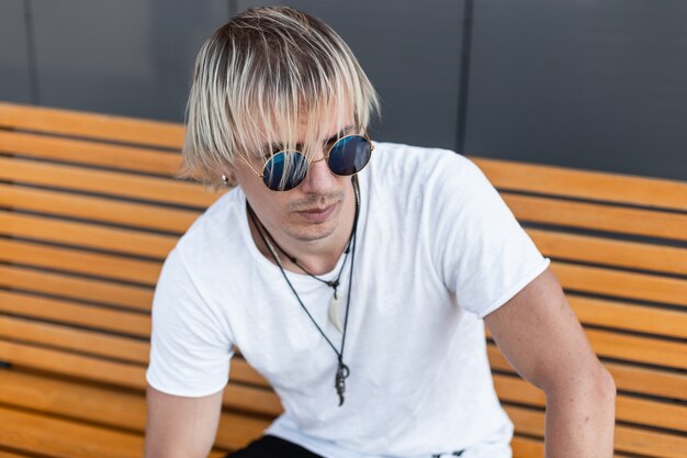 Handsome young man in trendy white t-shirt in stylish blue sunglasses with amulets on a neck is sitting on a wooden bench outdoors. American city guy is blond relaxes in the city. Street style