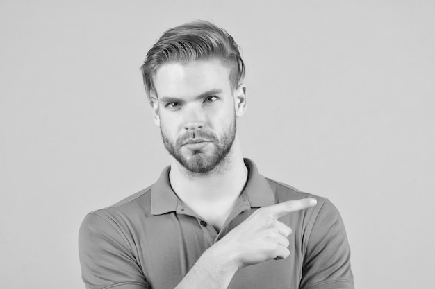 Handsome young man in tshirt looking at camera and pointing away while standing against grey background