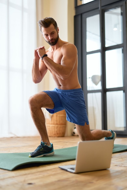 Handsome young man using laptop and doing lunges at home