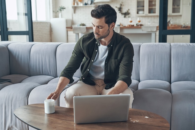 Handsome young man using laptop and drinking coffee while sitting on the sofa at home