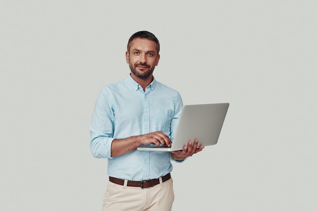 Bel giovane che usa il computer portatile e sorride mentre sta in piedi su uno sfondo grigio