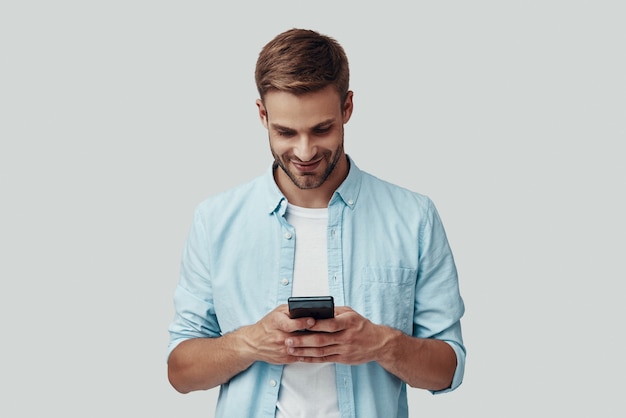 Handsome young man using smart phone and smiling while standing against grey background