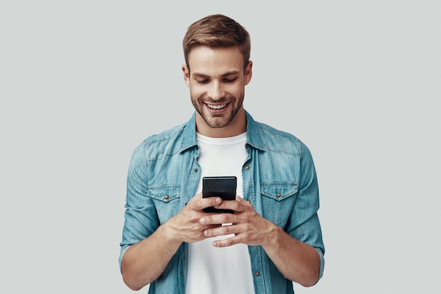 Photo handsome young man using smart phone and smiling while standing against grey background