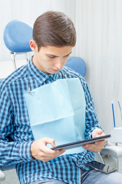 歯科医院を訪問中のハンサムな若い男。彼は椅子に座って、タブレットを手に持って歯科医を待っています。歯科
