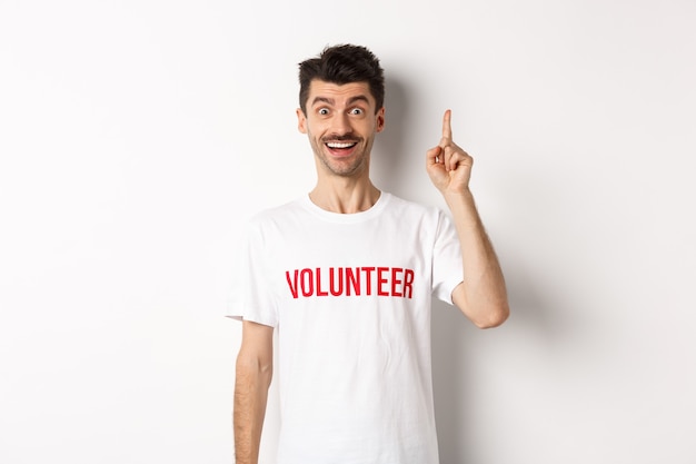 Handsome young man in volunteer t-shirt having an idea, raising finger and saying suggestion, pointing up, standing over white.