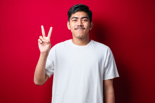 Handsome young man wearing white clothes showing and pointing up with fingers number two while smiling confident and happy over red background