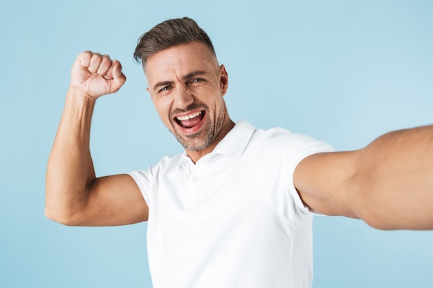 Handsome young man wearing white t-shirt standing over blue, taking a selfie