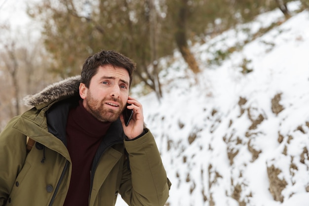 Handsome young man wearing winter jacket standing outdoors, talking on mobile phone