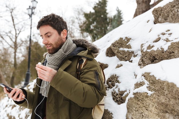 Handsome young man wearing winter jacket using mobile phone while walking outdoors, listening to music with earphones