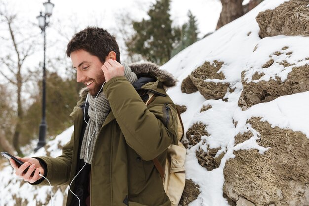 Handsome young man wearing winter jacket using mobile phone while walking outdoors, listening to music with earphones