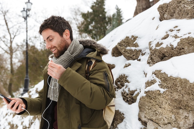 Handsome young man wearing winter jacket using mobile phone while walking outdoors, listening to music with earphones