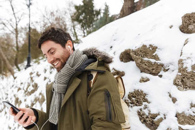 handsome young man wearing winter jacket using mobile phone while walking outdoors, listening to music with earphones