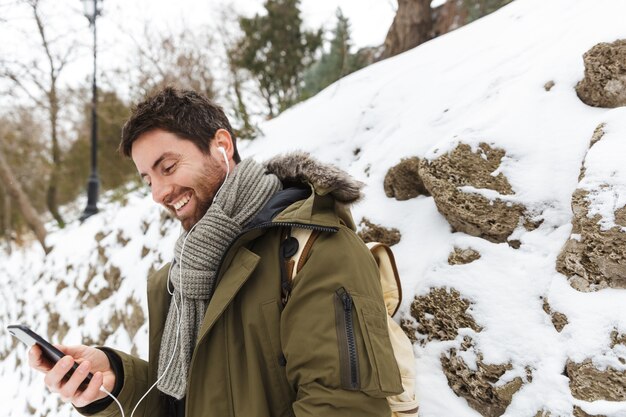 Handsome young man wearing winter jacket using mobile phone while walking outdoors, listening to music with earphones