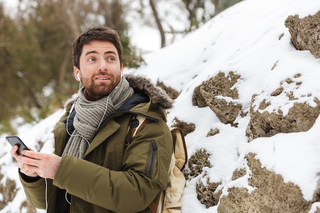 Handsome young man wearing winter jacket using mobile phone while walking outdoors, listening to music with earphones