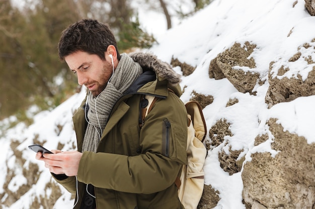 handsome young man wearing winter jacket using mobile phone while walking outdoors, listening to music with earphones