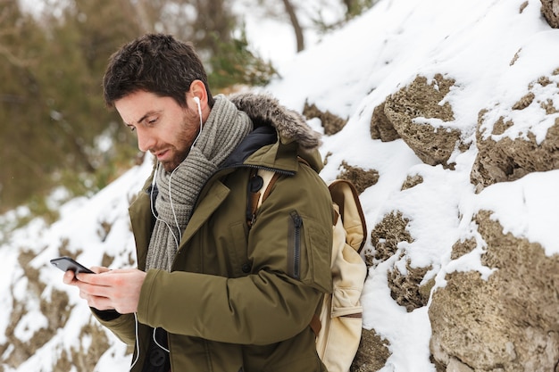 Handsome young man wearing winter jacket using mobile phone while walking outdoors, listening to music with earphones