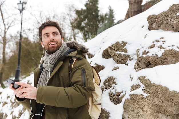 Handsome young man wearing winter jacket using mobile phone while walking outdoors