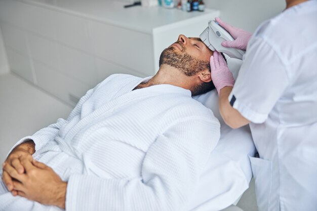 Handsome young man in white bathrobe lying on daybed while receiving laser facial treatment in cosmetological clinic