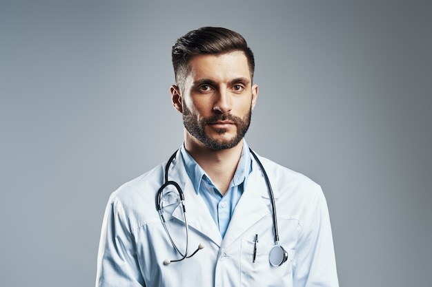 Handsome young man in white lab coat looking at camera while standing against grey wall