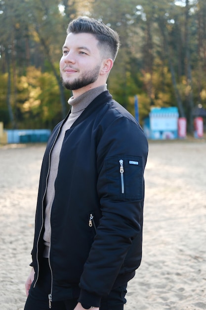 Handsome young man with a beard and in a black jacket posing on the beach