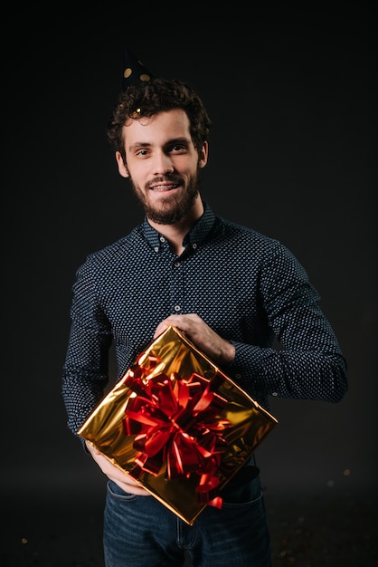Handsome young man with beard in a festive cap holds a holiday box with a gift