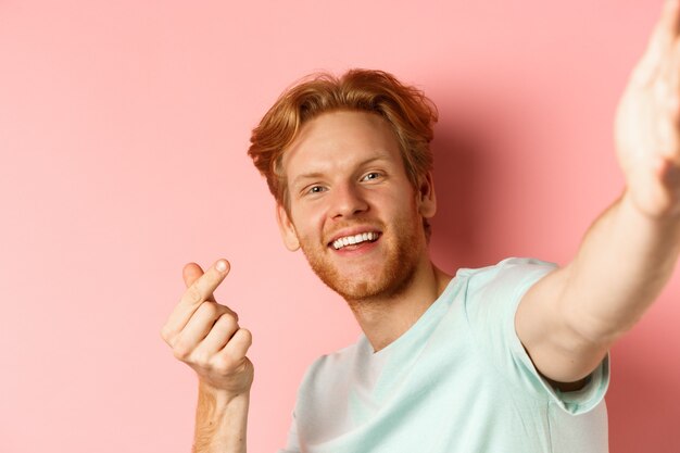 Handsome young man with beard and red hair taking selfie against pink background, stretch out hand to hold smartphone camera and showing korean heart gesture, pink background.