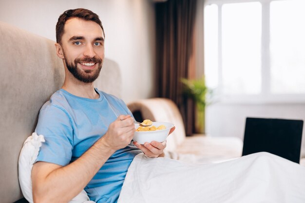 Handsome young man with beard smiling eating breakfast in bed