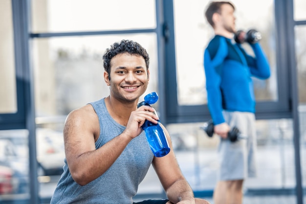 Handsome young man with bottle of water resting at gym other man workout on blurred background