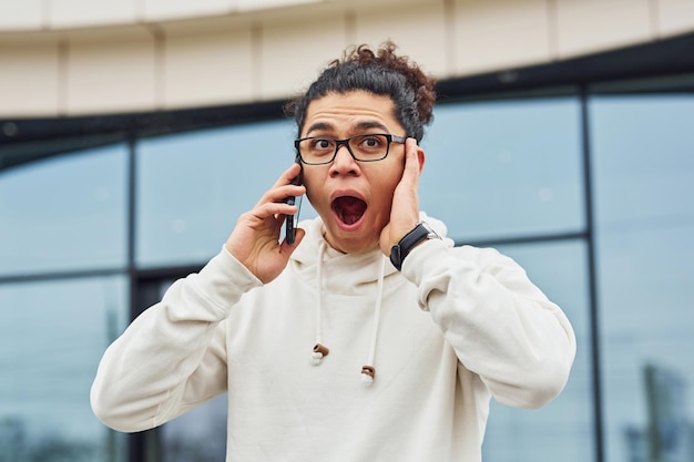 Photo handsome young man with curly black hair is on the street against building talking by the phone.